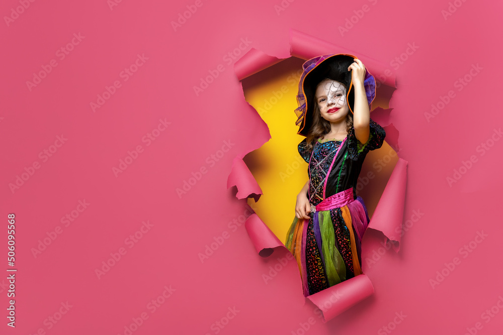 Fototapeta premium Laughing funny child girl in a witch costume of halloween looking, smiling and scares through a hole of red, yellow paper background. Copyspace