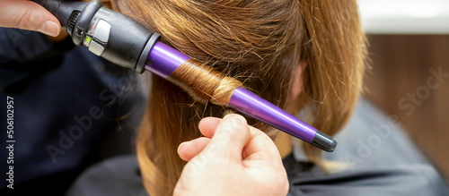 The hairstylist makes curls hairstyle of long brown hair with the curling iron in hairdresser salon, close up