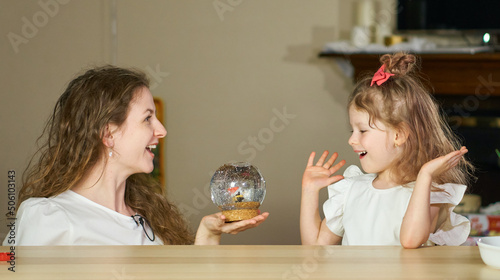 Magic Surprise for child. Snow globe as gift. Happy childhood. Woman surprised little beautiful girl in white. Happy childhood. Have fun together. Give gift to children. Mom congratulated her daughter