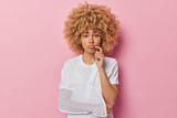 People recovery and rehabilitation concept. Dissatisfied curly haired European woman looks unhappily at camera has broken arm after accident wears white t shirt isolated over pink background.
