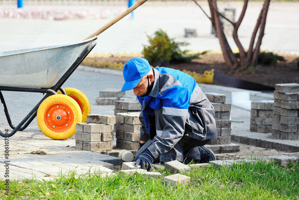 Bricklayer in work clothes sits on sidewalk and lays out paving slabs ...