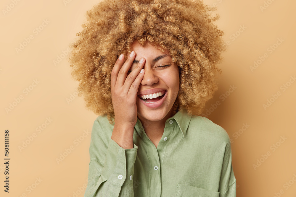 Happy cheerful woman makes face palm laughs joyfully expresses sincere feelings and positive emotions dressed in casual linen shirt isolated over brown background. Peope and happiness concept - obrazy, fototapety, plakaty 