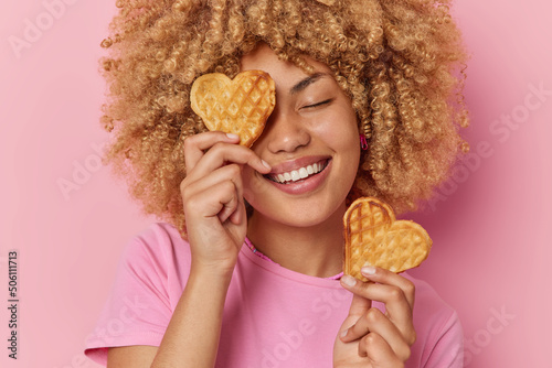 Positive woman with curly hair holds two heart shaped waffles baked by herself smiles broadly keeps eyes closed dressed in casual t shirt isolated over pink background. Delicious food concept