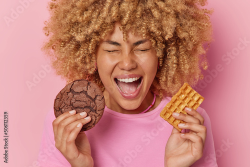 Overjoyed curly haired young woman holds delicious chocolate cookie and homemade waffle has sweet tooth has addiction to sugar exclaims loudly keeps mouth opened isolated over pink background
