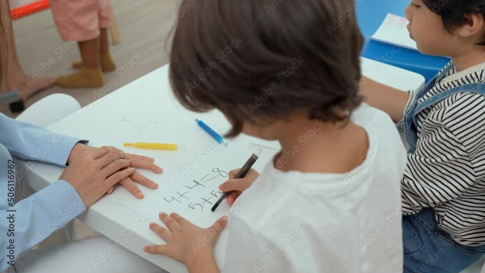 Asian female teacher teach and write on whiteboard while children sit on the chair in classroom, Multi-ethnic preschool teacher and students in classroom. Kindergarten pre school concept. Asian female teacher teach and write on whiteboard while children sit on the chair in classroom, Multi-ethnic preschool teacher and students in classroom. Kindergarten pre school concept.