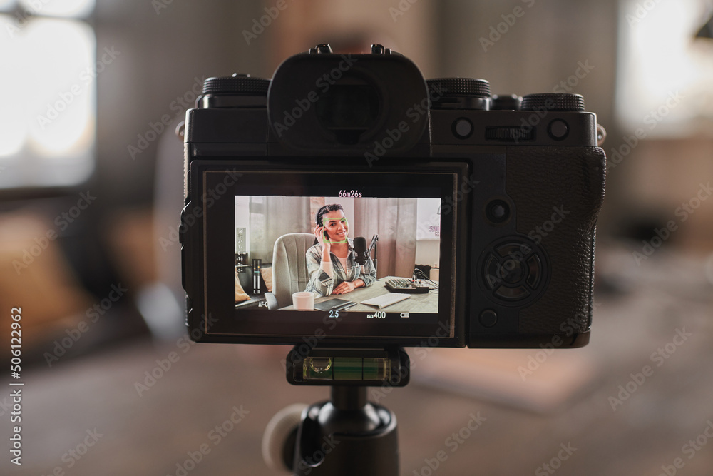 Horizontal close-up shot of camera recording video of stylish woman wearing headphones sitting at desk in loft room creating content for her blog