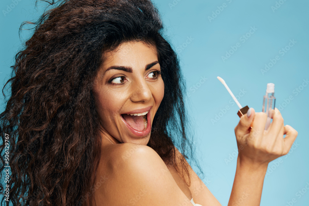 Obraz premium Excited happy attractive curly Latin lady look at lip gloss lipstick posing isolated over pastel blue background. Cosmetic lips product ad Natural beauty Makeup concept Studio portrait Copy space