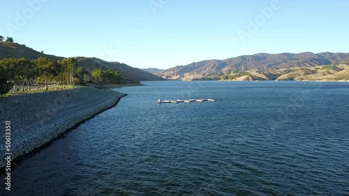 aerial footage of a gorgeous vast blue rippling lake surrounded by majestic mountain ranges with lush green trees and plants on the banks and a clear blue sky at Castaic Lake in Castaic California USA photo