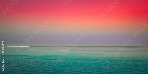 Red sunset clouds in the foggy sky, purple horizon, turquoise-colored seawater, and rolling waves