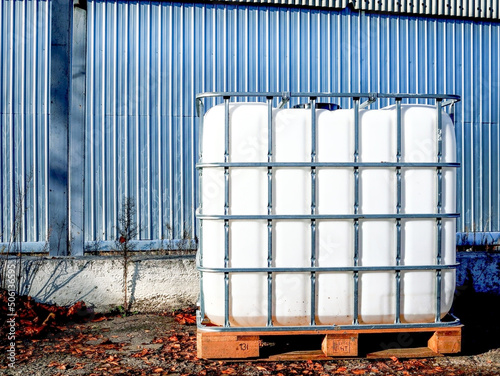 White square water tank with metal grate container for liquid standing outdoors on wooden pallet in front of a steel wall