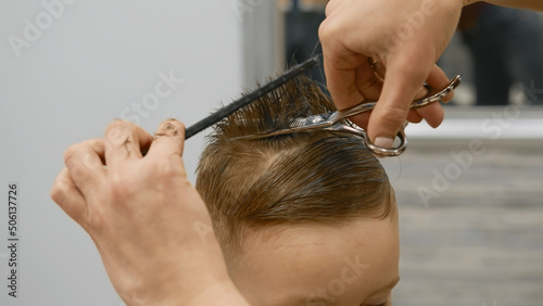 Process of hairdresser cutting hair. Blond caucasian boy is sitting in barber shop and hairdresser girl is cutting his hair. Child gets fashionable haircut, hairdresser cuts child hair with scissors.