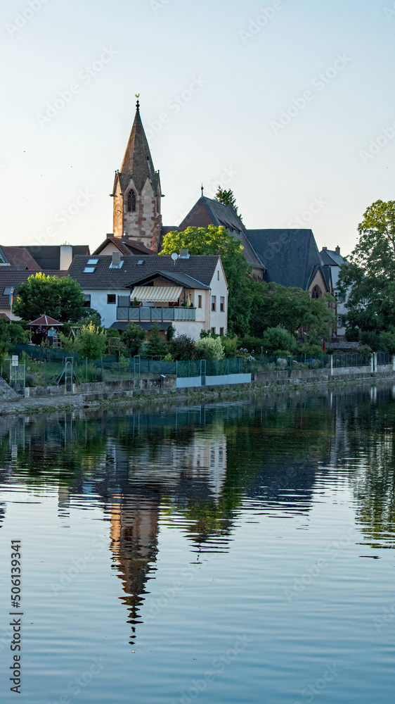 The church tower and other village buildings reflect in the placid waters of the Rhine River in the early evening.