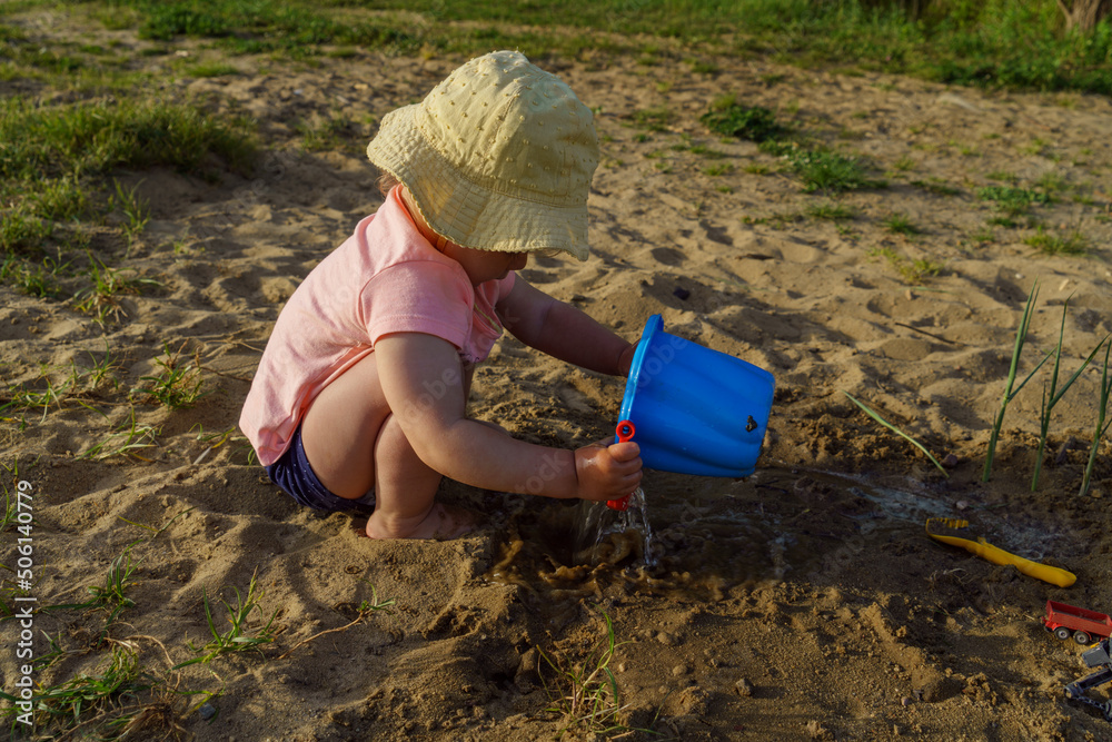 little baby playing with a blue plastic bucket filled with water on a background of sand, a child with bare feet pours water from a bucket learns something new fun childhood in the summer on vacatio little baby playing with a blue plastic bucket filled with water on a background of sand, a child with bare feet pours water from a bucket learns something new fun childhood in the summer on vacatio