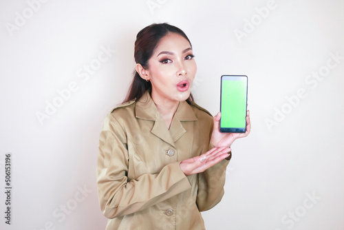 Shocked Asian woman wearing brown uniform pointing finger at green screen on smartphone in her hand photo