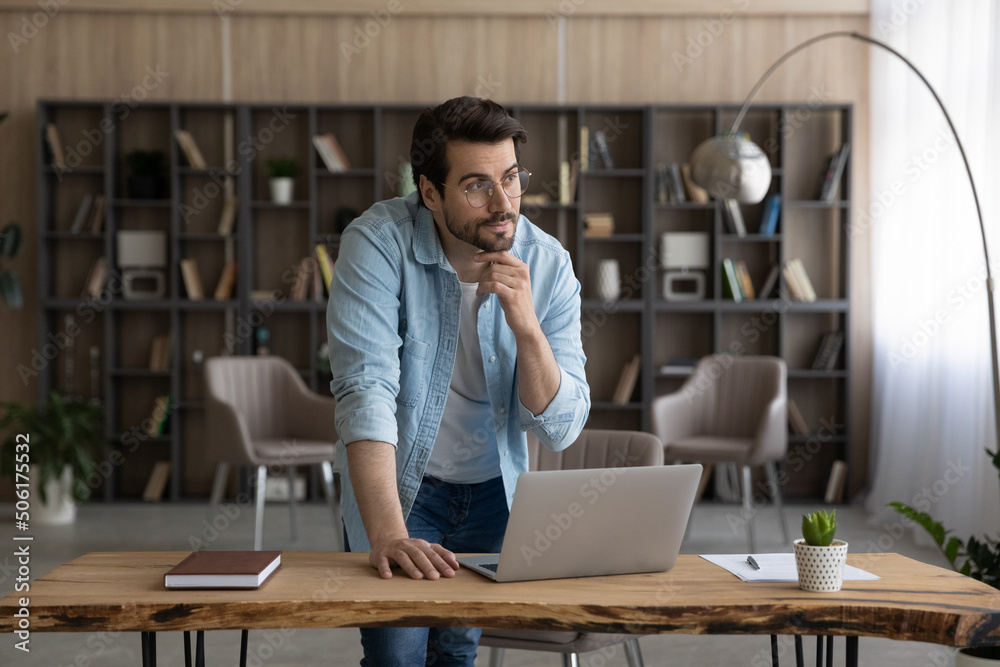 Work on idea. Thoughtful casual male in glasses stand by desk with pc look at distance touch chin create project ponder on business plan. Pensive young man scientist do research think on new solution Work on idea. Thoughtful casual male in glasses stand by desk with pc look at distance touch chin create project ponder on business plan. Pensive young man scientist do research think on new solution