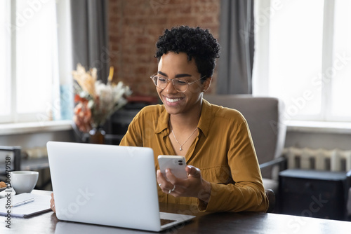 African woman sit at workplace desk holds cellphone staring at laptop, synchronize data between computer and gadget in office, use corporate devices and business application, plan work, use organizer