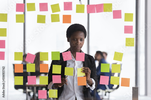 Serious African woman attaching sticky notes on transparent wall in boardroom, jotting corporate goals, develop ideas using colored post-it stickers Fototapet