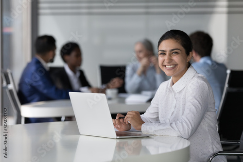 Indian businesswoman satisfied by company position, career growth at desk with laptop smile pose look at camera, diverse colleagues on background. Successful promoted company employee portrait concept