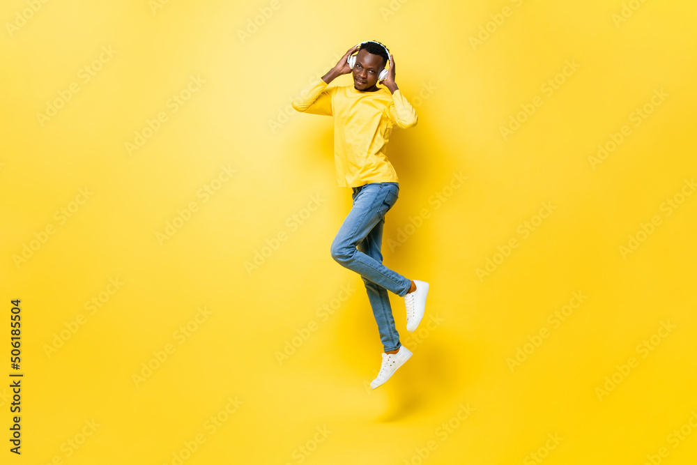 Young African man listening to music on headphones and jumping in isolated yellow studio background
