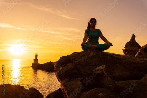 A woman doing meditation and yoga exercises at sunset next to a lighthouse in the sea  healthy and naturist life  outdoor pilates  ardha padmasana