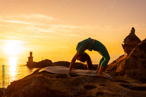 Urdhva Dhanurasana, a woman doing meditation and yoga exercises on a rock at sunset next to a lighthouse in the sea, healthy and naturist life