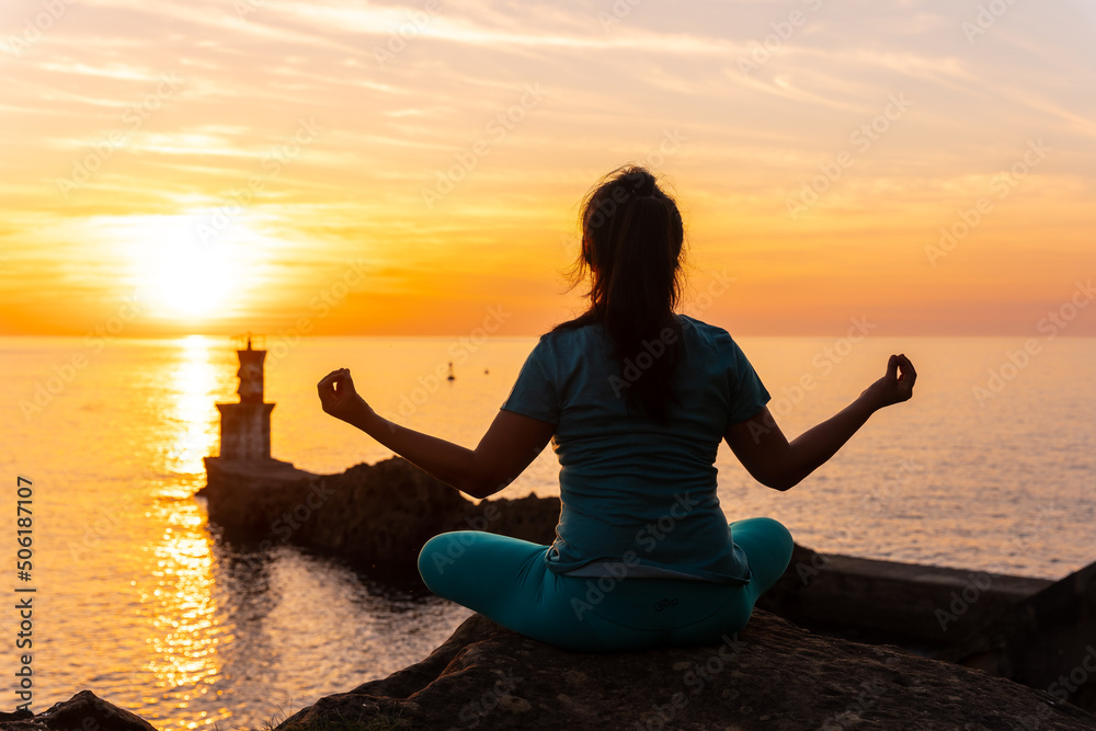 A woman doing meditation on a rock at sunset next to a lighthouse in the sea, healthy and naturist life
