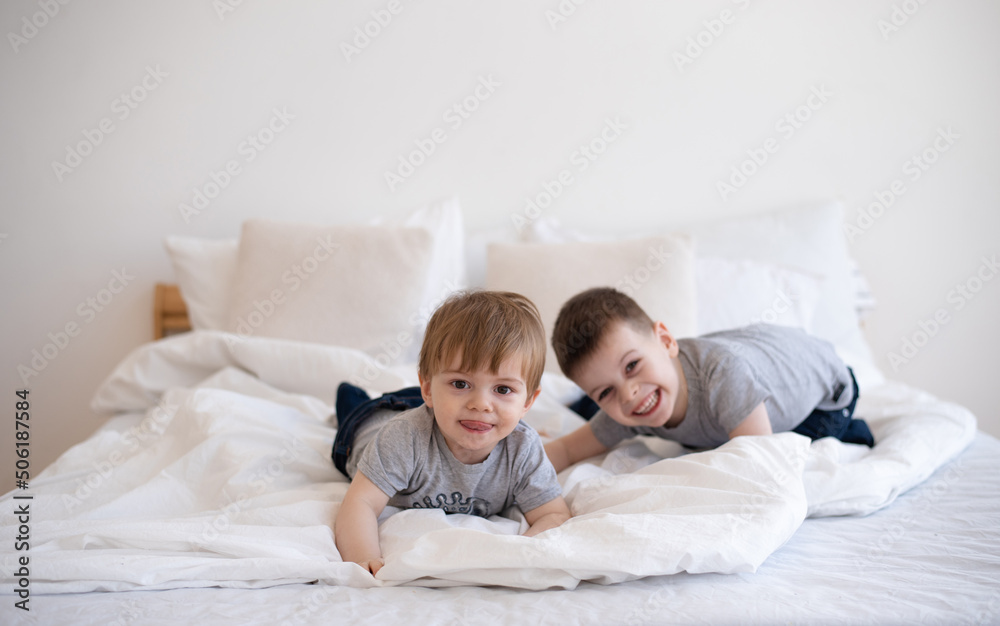 Smiling two boys in gray t-shirts lie and playing on white blanket on bed. Cheerful and happy childhood in family. Exercise and play good for health. Together joyful and interesting. Smiling two boys in gray t-shirts lie and playing on white blanket on bed. Cheerful and happy childhood in family. Exercise and play good for health. Together joyful and interesting.