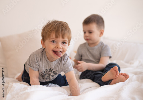 Smiling two boys in gray t-shirts sit and playing on white blanket on bed. Cheerful and happy childhood in family.  Exercise and play good for health. Together joyful and interesting. 