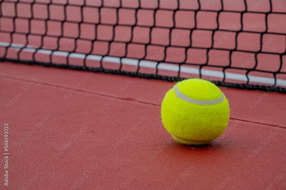 Tennis ball next to the net of a red hard surface tennis court.