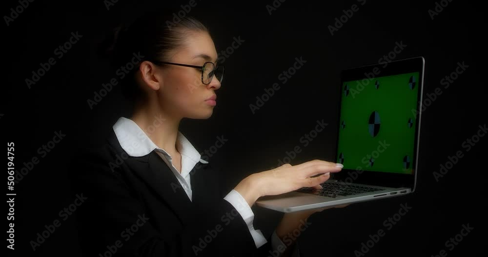 Thoughtful business woman with glasses and office clothes is typing on a laptop with a green screen for copy space, thinking standing on a black background. Chroma key mock-up Thoughtful business woman with glasses and office clothes is typing on a laptop with a green screen for copy space, thinking standing on a black background. Chroma key mock-up