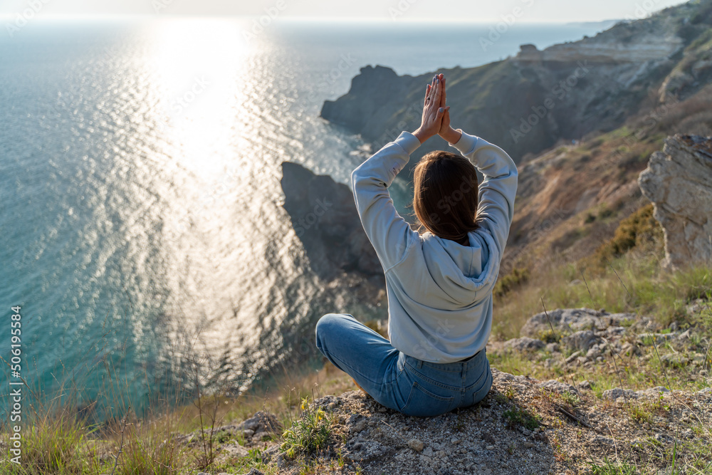Woman tourist enjoying the sunset over the sea mountain landscape. Sits outdoors on a rock above the sea. She is wearing jeans and a blue hoodie. Healthy lifestyle, harmony and meditation Woman tourist enjoying the sunset over the sea mountain landscape. Sits outdoors on a rock above the sea. She is wearing jeans and a blue hoodie. Healthy lifestyle, harmony and meditation