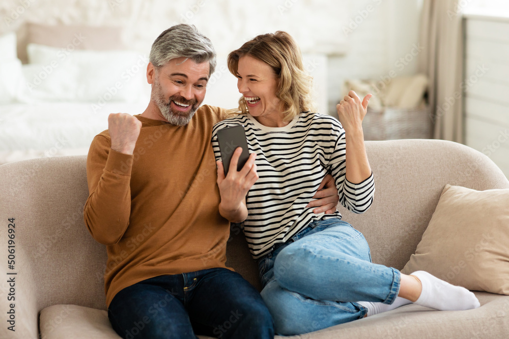 Happy Middle Aged Couple Using Smartphone Shaking Fists At Home Happy Middle Aged Couple Using Smartphone Shaking Fists At Home