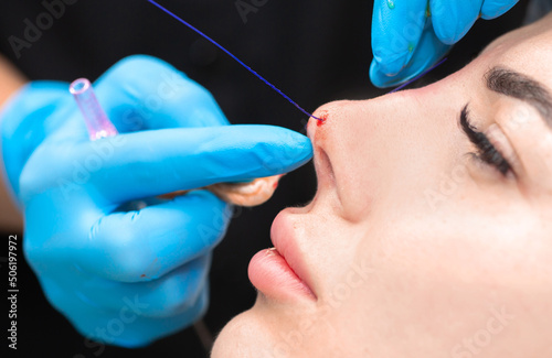Fototapeta Naklejka Na Ścianę i Meble -  Cosmetic procedure for non-surgical rhinoplasty. The beautician inserts cosmetic threads into the nose with a needle to reshape the nose, close-up. Plastic surgery, cosmetology