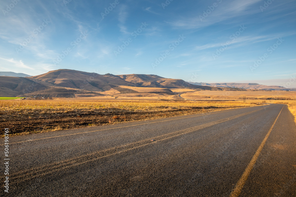 Fototapeta premium A tarmac road cuts through the dry, arid landscape of Kwazulu Natal near Drakensberg, South Africa.