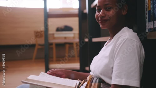 A schoolgirl sitting in front of library's bookshelf and reading a book preparing for an exam.