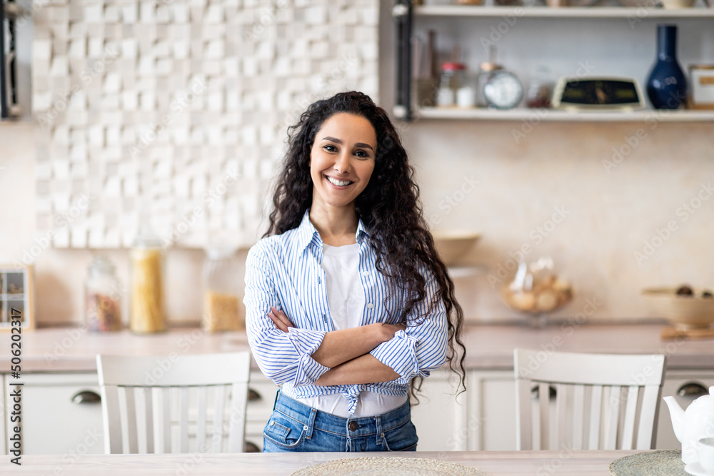 Portrait of happy latin housewife posing with folded arms in kitchen, smiling and looking at camera, free space
