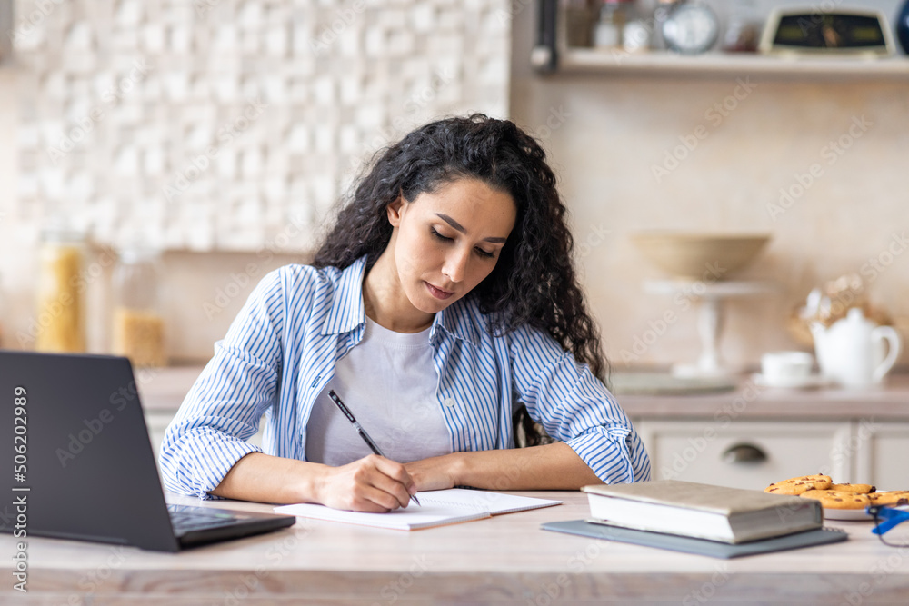 Young latin businesswoman writing at notepad while sitting in front of brand new laptop at kitchen interior Young latin businesswoman writing at notepad while sitting in front of brand new laptop at kitchen interior