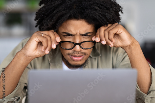 Concentrated curly young black male in glasses looks at laptop screen at table in cafe interior, close up photo