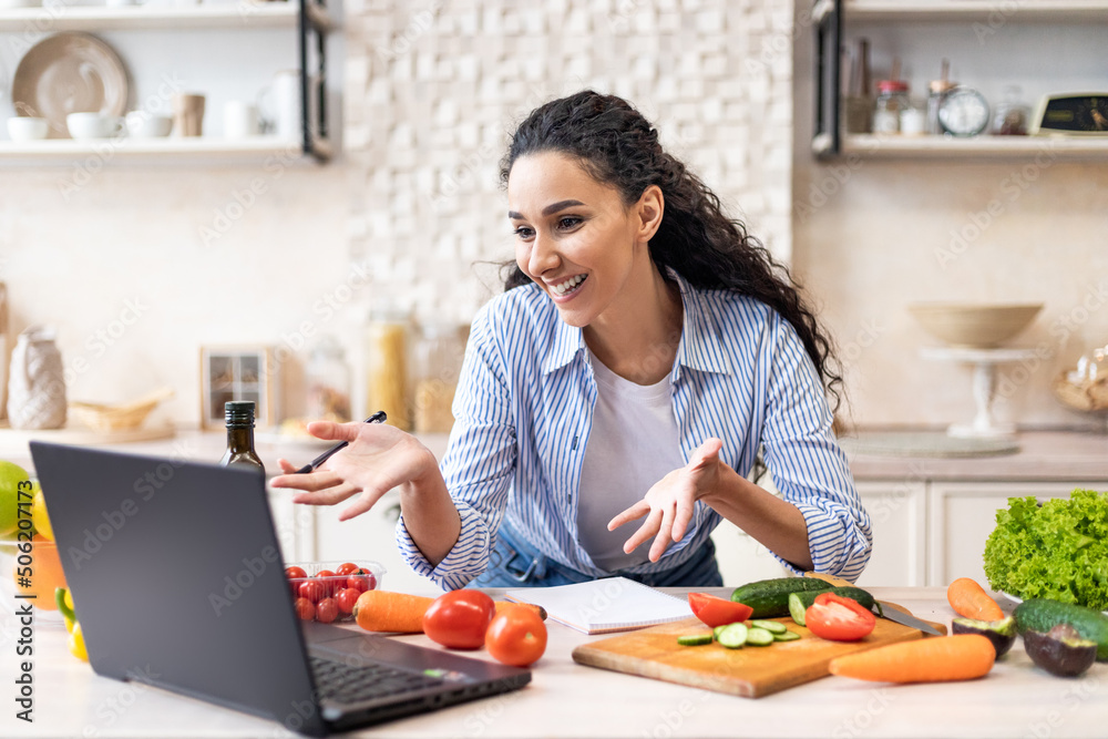 Happy latin woman talking to laptop webcamera while preparing fresh salad in kitchen interior, having video call
