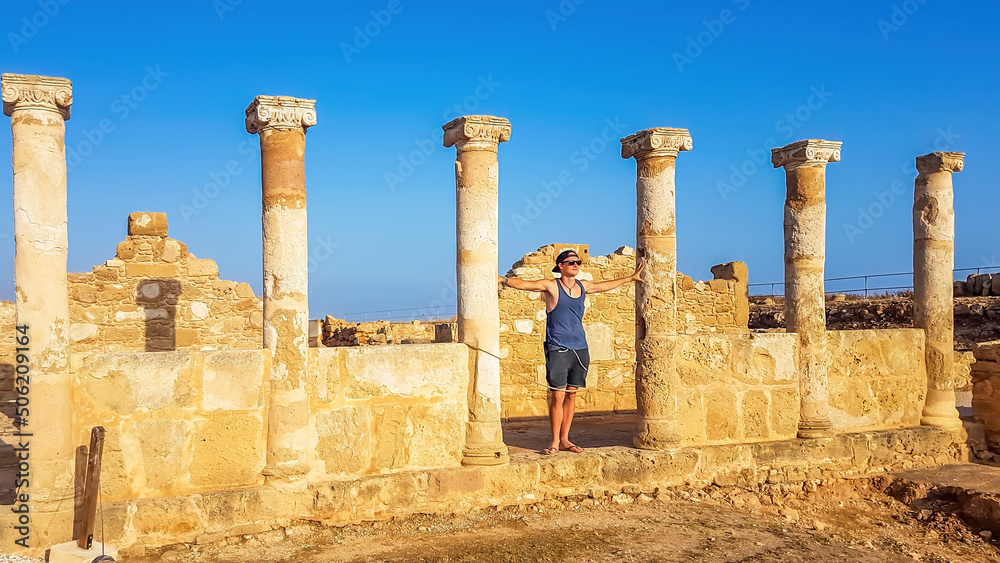 Young man standing between the ruins of an ancient temple, what is left are temple columns, Kato Paphos Archeological Park. Sandy color of the construction. Clear and blue sky.