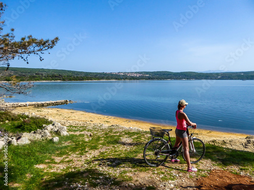 A girl in a sporty outfit stands on her bike by the seashore and admires the view in front of her. Coast is partially sandy and partially stony and grassy. The water in the bay is calm and clean photo