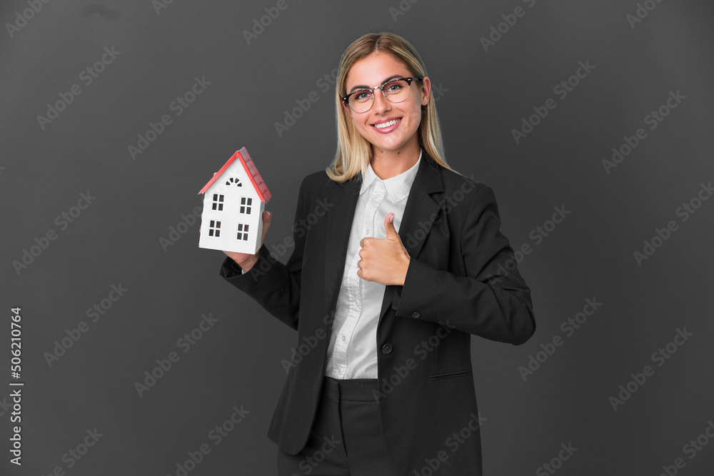 Blonde Uruguayan girl holding a house toy isolated on black background giving a thumbs up gesture Blonde Uruguayan girl holding a house toy isolated on black background giving a thumbs up gesture