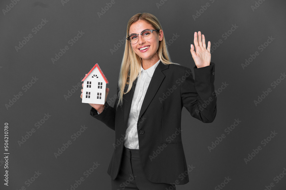 Blonde Uruguayan girl holding a house toy isolated on black background saluting with hand with happy expression Blonde Uruguayan girl holding a house toy isolated on black background saluting with hand with happy expression