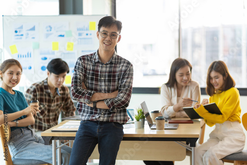 Portrait of happy smiling young businessman in office meeting room looking at camera while colleagues are meeting in the background.