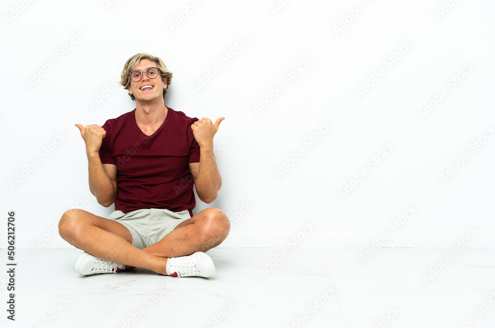 Young English man sitting on the floor with thumbs up gesture and smiling