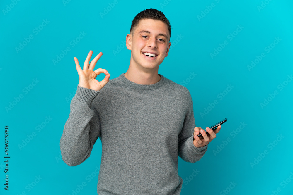 Young caucasian man isolated on blue background using mobile phone and doing OK sign Young caucasian man isolated on blue background using mobile phone and doing OK sign