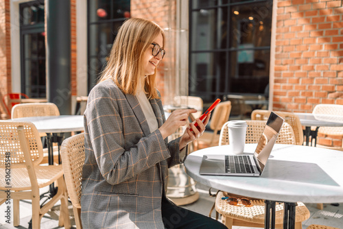 Smiling woman sitting in cafeteria holding coffee mug and working on laptop. Businesswoman checking email on smartphone. Beautiful 30s woman and using laptop at cafe while drinking a cup of tea. 