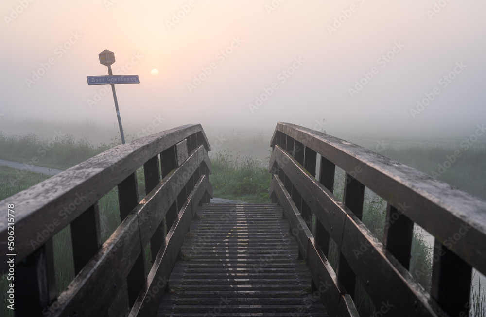 Small wooden bridge in a nature area during a foggy, spring sunrise. The sign is the street name of the path: Boer Goensepad.