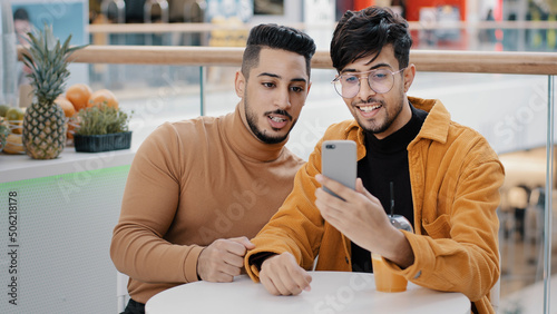 Two happy arab friend bloggers sitting in cafe recording video blog on phone communicating via webcam looking at smartphone screen take selfie making videos on social media using mobile application