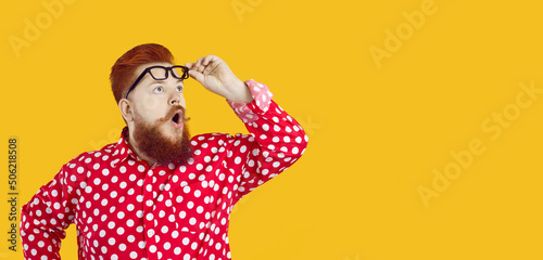 Funny charismatic chubby man with shocked expression looks at copy space on yellow background. Red-bearded fat man in stylish red shirt in white peas looks amazed raising his glasses from his eyes.